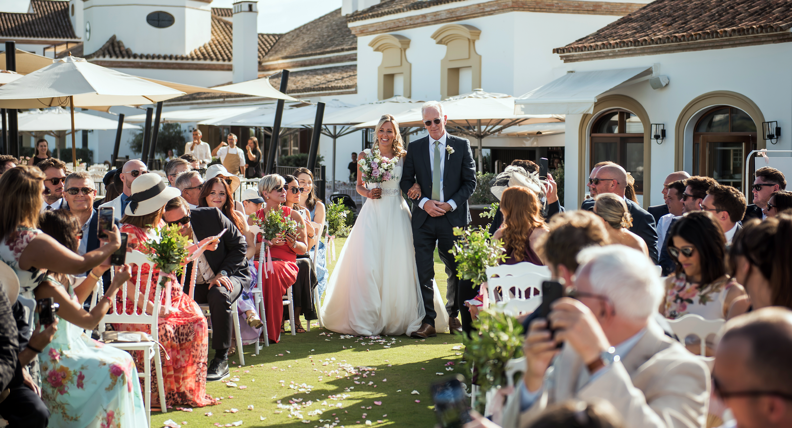 A bride in a white dress walks down the aisle with an older man at an outdoor wedding ceremony, surrounded by seated guests in formal attire.