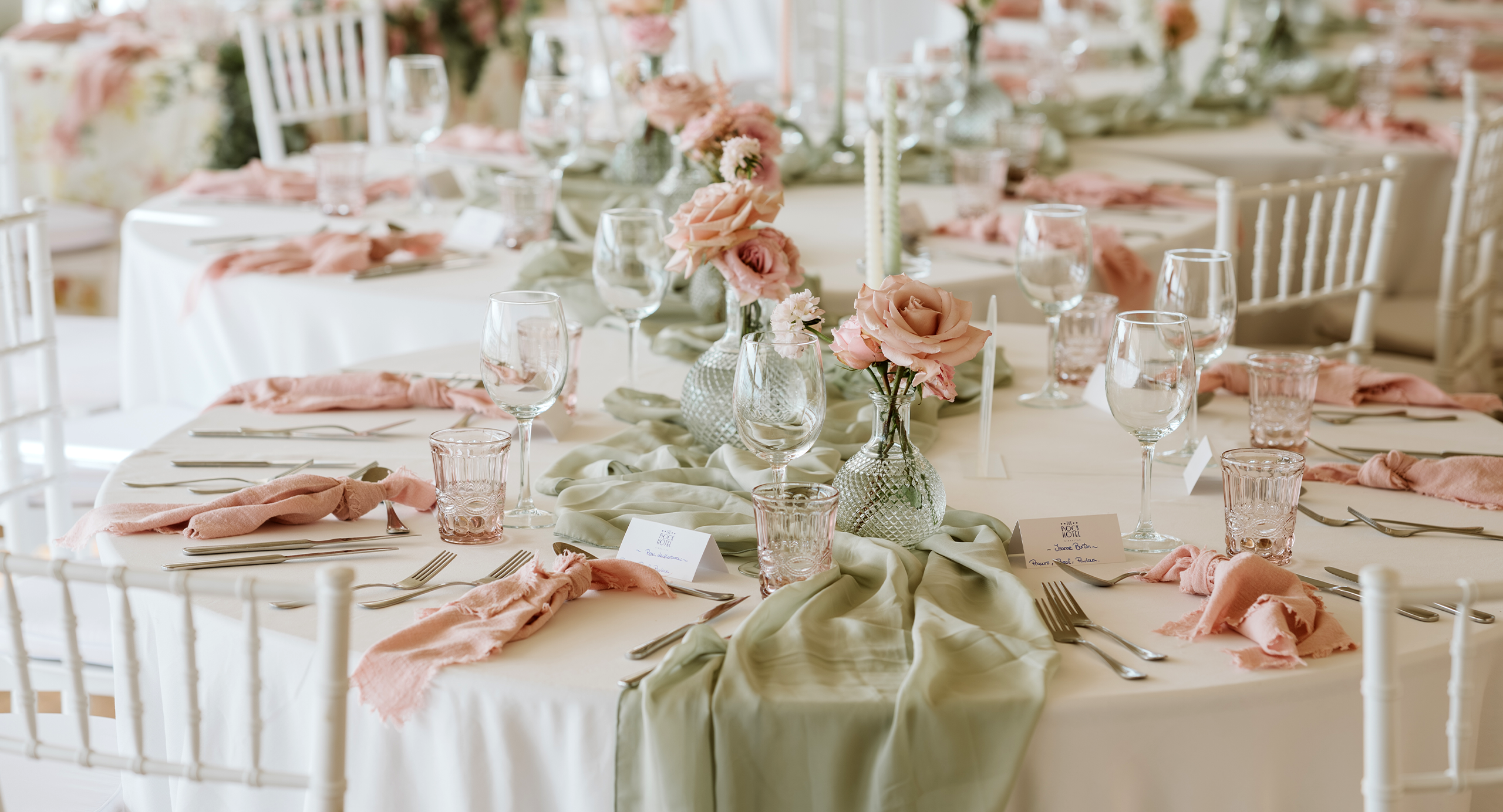 Round tables set for an event with glassware, cutlery, sage green table runners, pink napkins, and floral centerpieces with roses. White chairs surround the tables.