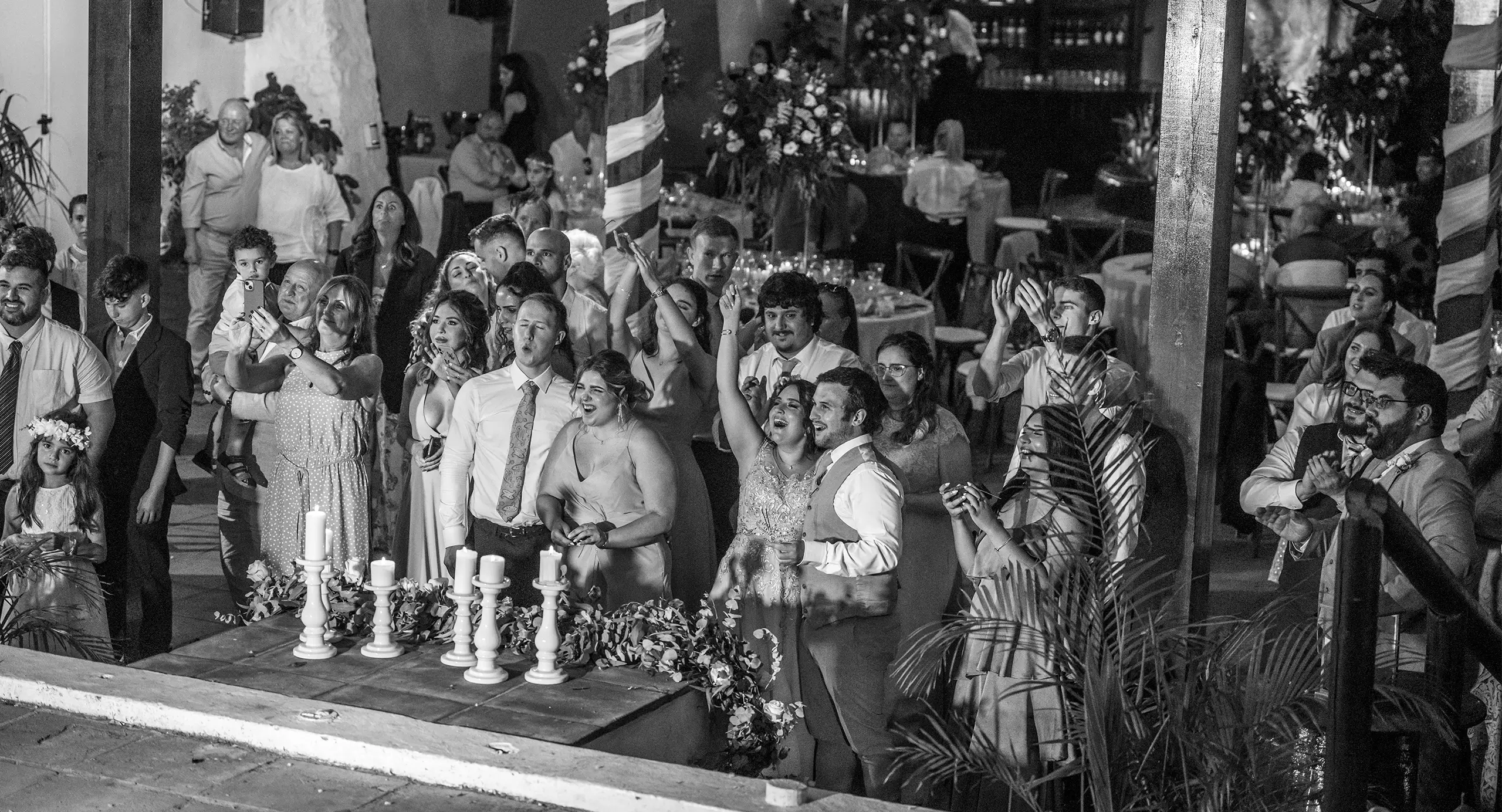 A large group of people dressed formally gathers indoors, watching something off-camera with excited and joyful expressions near a decorated table with candles.