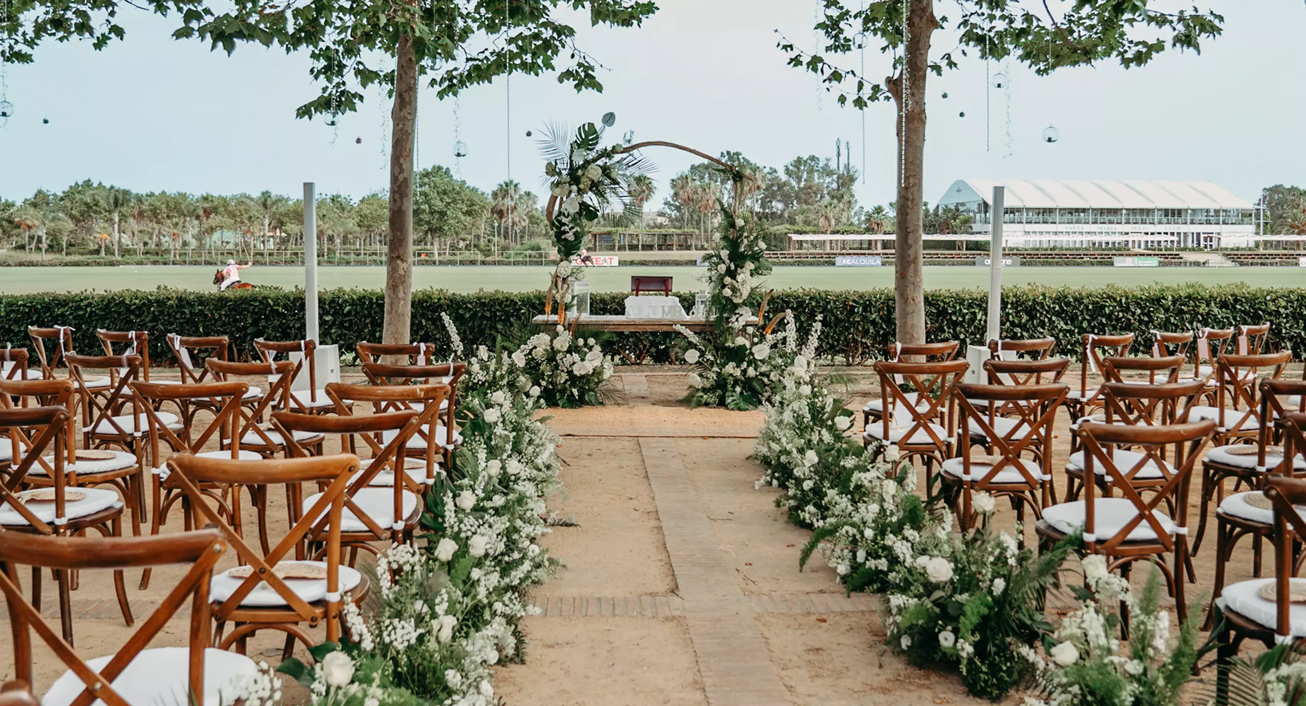 Outdoor wedding ceremony setup with wooden chairs, white floral arrangements along the aisle, and a decorated arch at the front under trees.