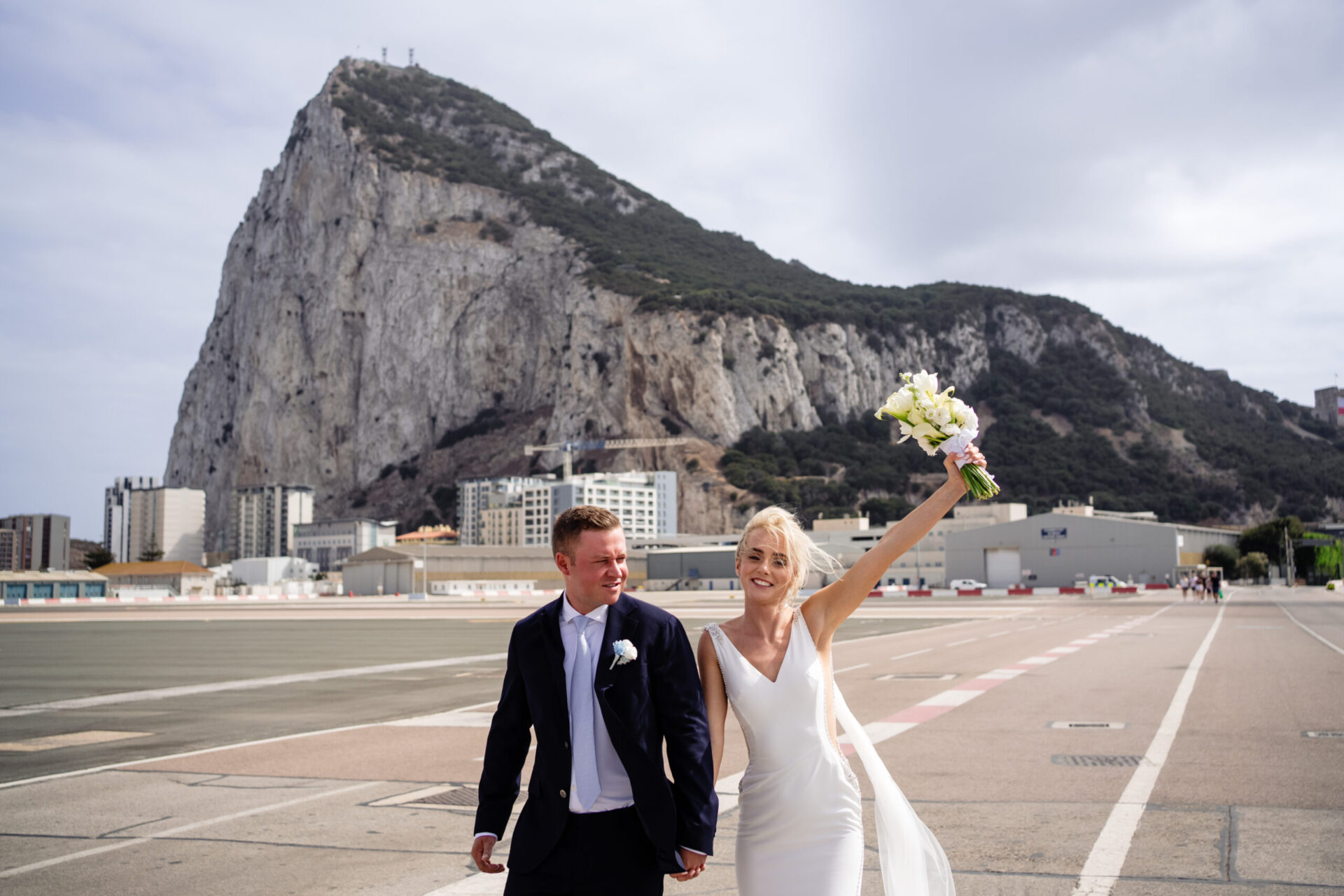 A bride and groom walk hand in hand, smiling, as the bride holds up her bouquet. The Rock of Gibraltar rises in the background under a partly cloudy sky.