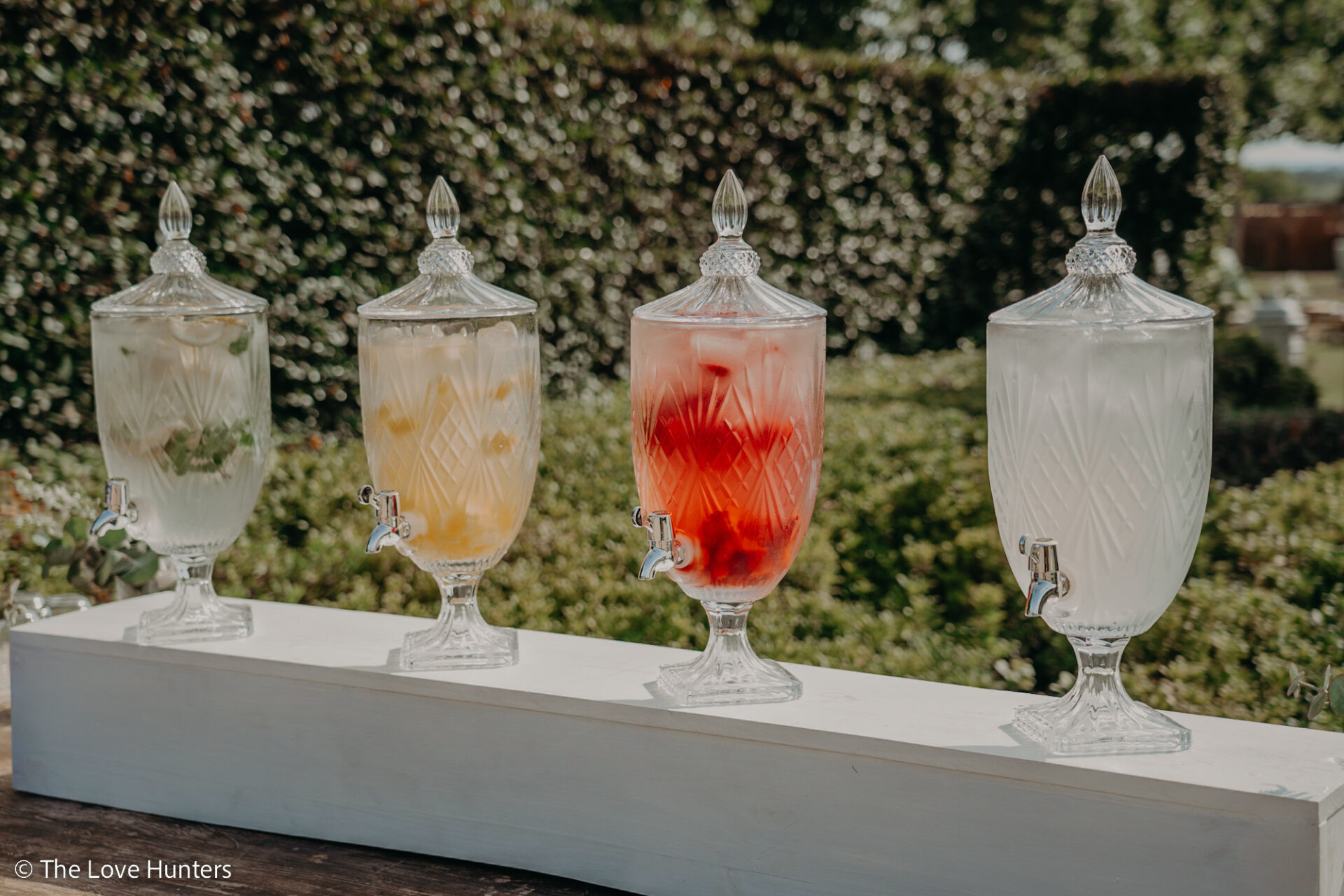 Four ornate glass drink dispensers filled with different fruit-infused waters are arranged in a row outdoors, each with a spigot, set against a green leafy garden background.