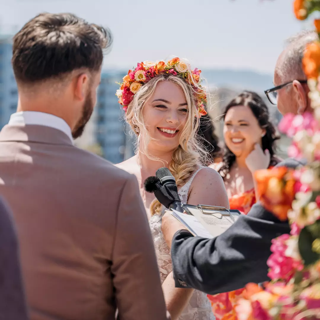 A bride wearing a floral crown smiles during an outdoor wedding ceremony, standing opposite the groom while an officiant holds a microphone.