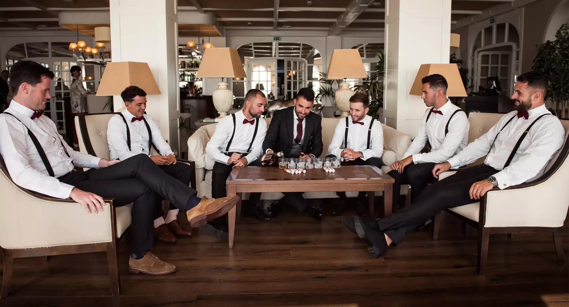 Seven men dressed in formal attire sit around a coffee table in a well-lit lounge, appearing to talk and prepare for an event.