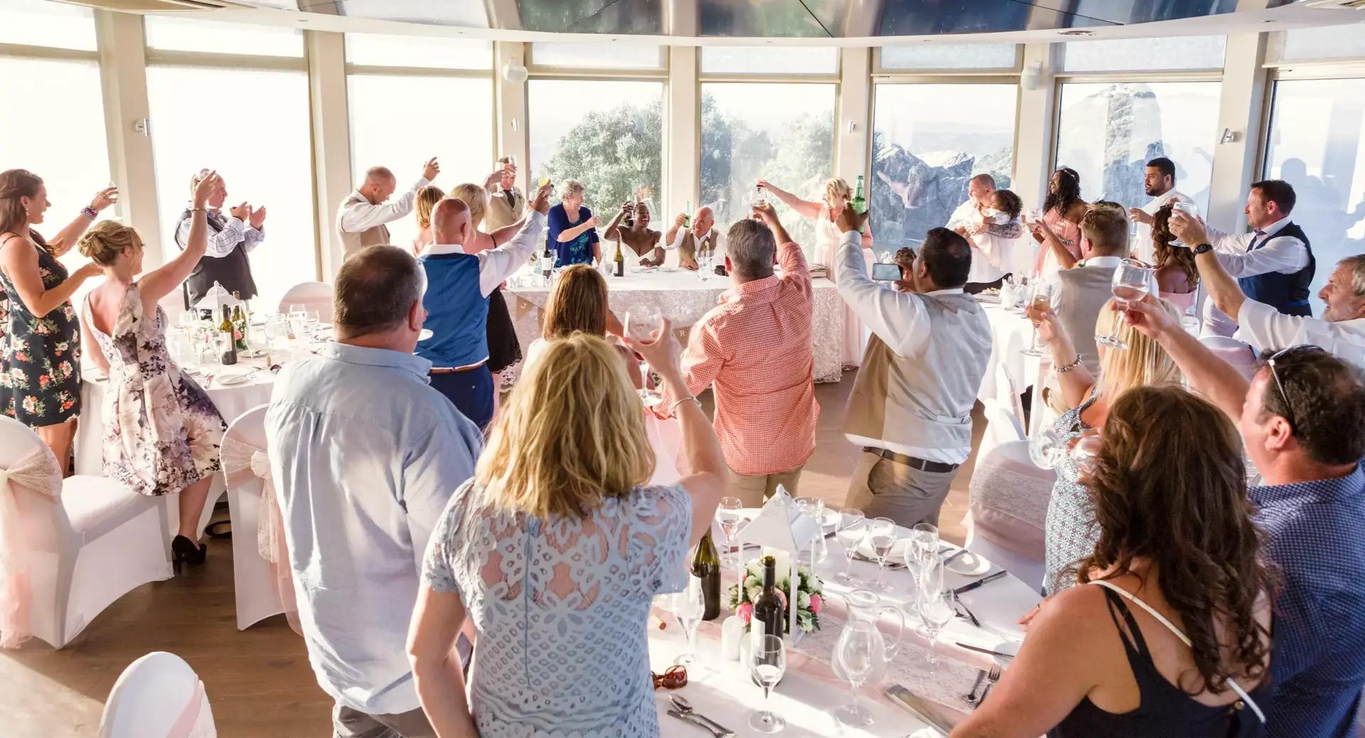 A group of people at a formal event raise their glasses in a toast while seated around decorated tables in a bright, windowed room.