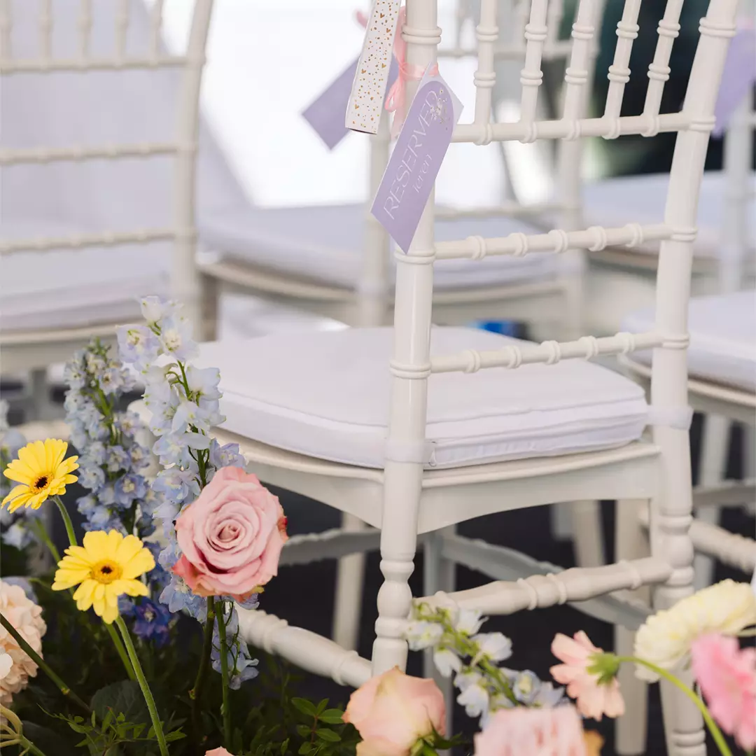 White chairs with padded seats are lined up, each with a name tag. Colorful flowers, including roses and daisies, are arranged at the base of the chairs.