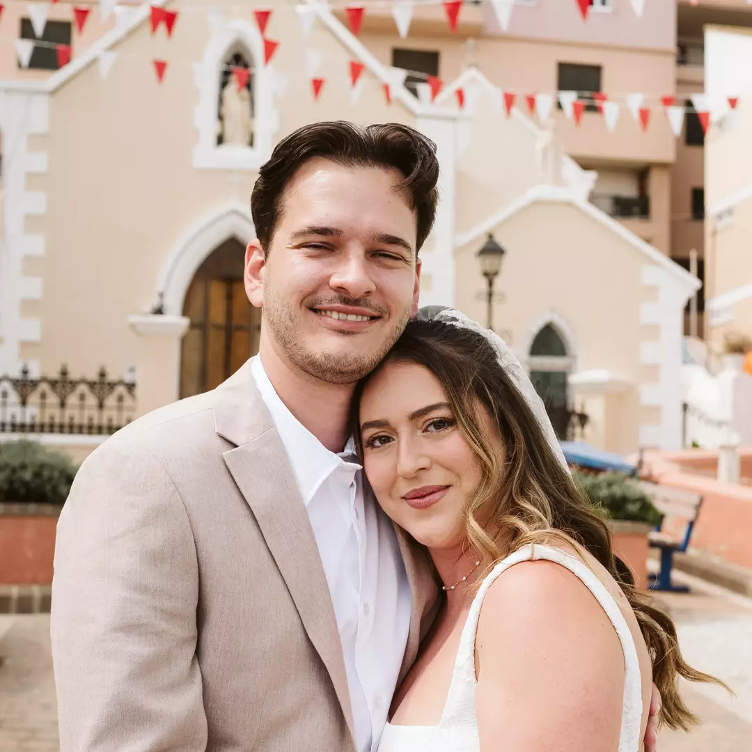 A couple in wedding attire poses closely together and smiles in front of a church decorated with red and white bunting.