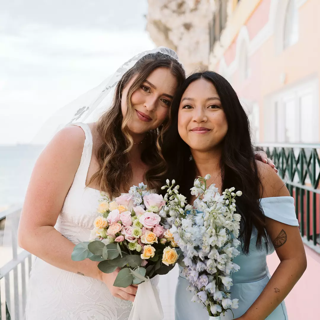 Two women stand side by side outdoors, smiling at the camera. One wears a wedding dress and holds a bouquet; the other wears a light blue dress and also holds flowers.