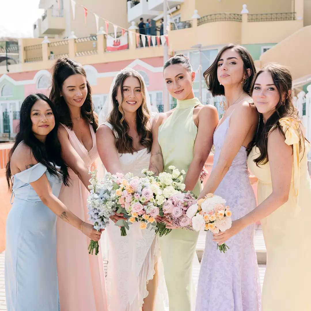 A bride in a white dress stands with five bridesmaids in pastel dresses, each holding bouquets, posing outdoors at a wedding venue.