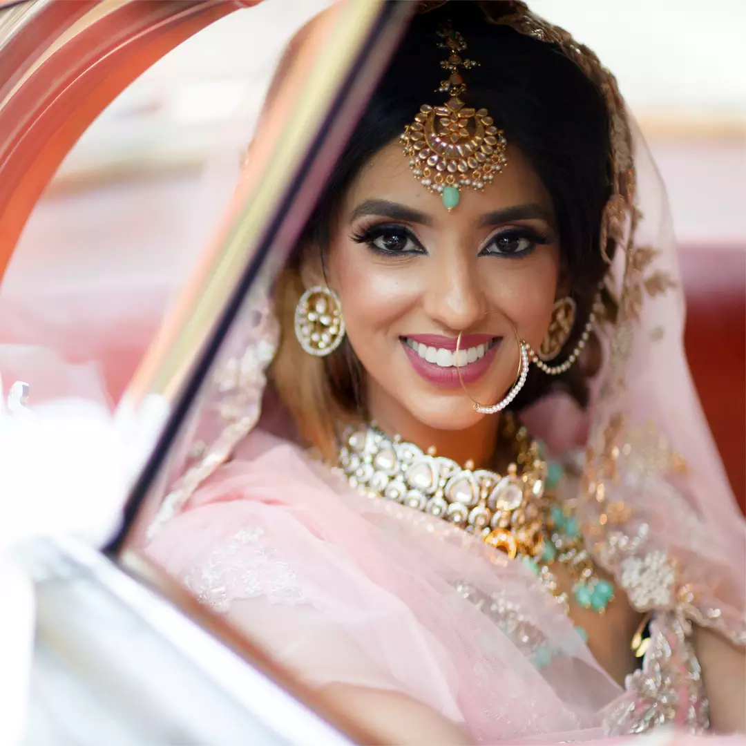A woman in traditional bridal attire, adorned with jewelry and a veil, smiles while seated in a car.