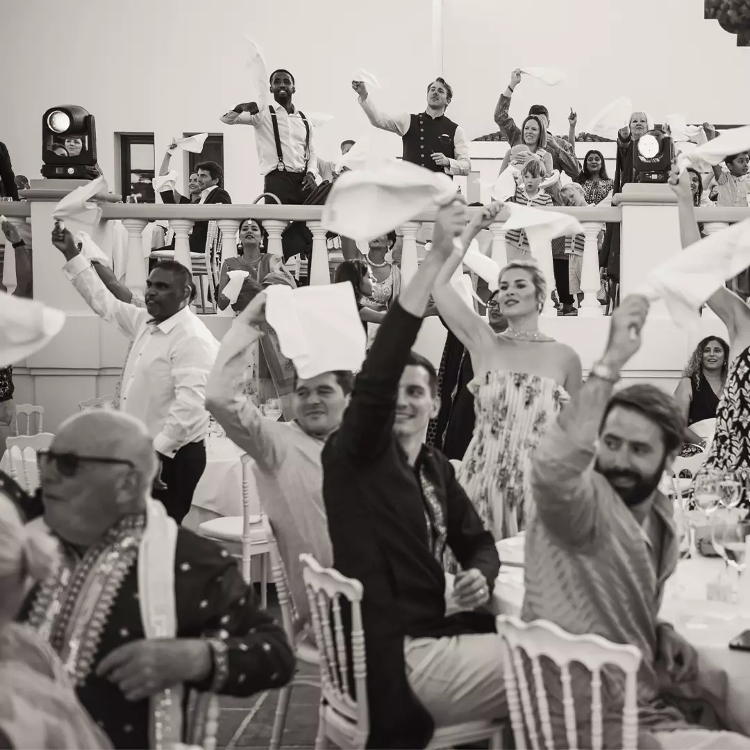 A group of people at a formal event or reception wave napkins in the air, some standing and others seated at round tables, in a lively atmosphere.