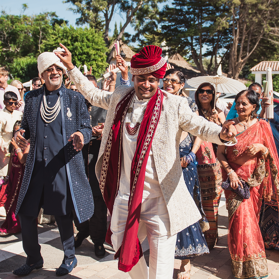 A groom in traditional Indian attire dances joyfully outdoors, surrounded by smiling guests in festive clothing during a wedding celebration.