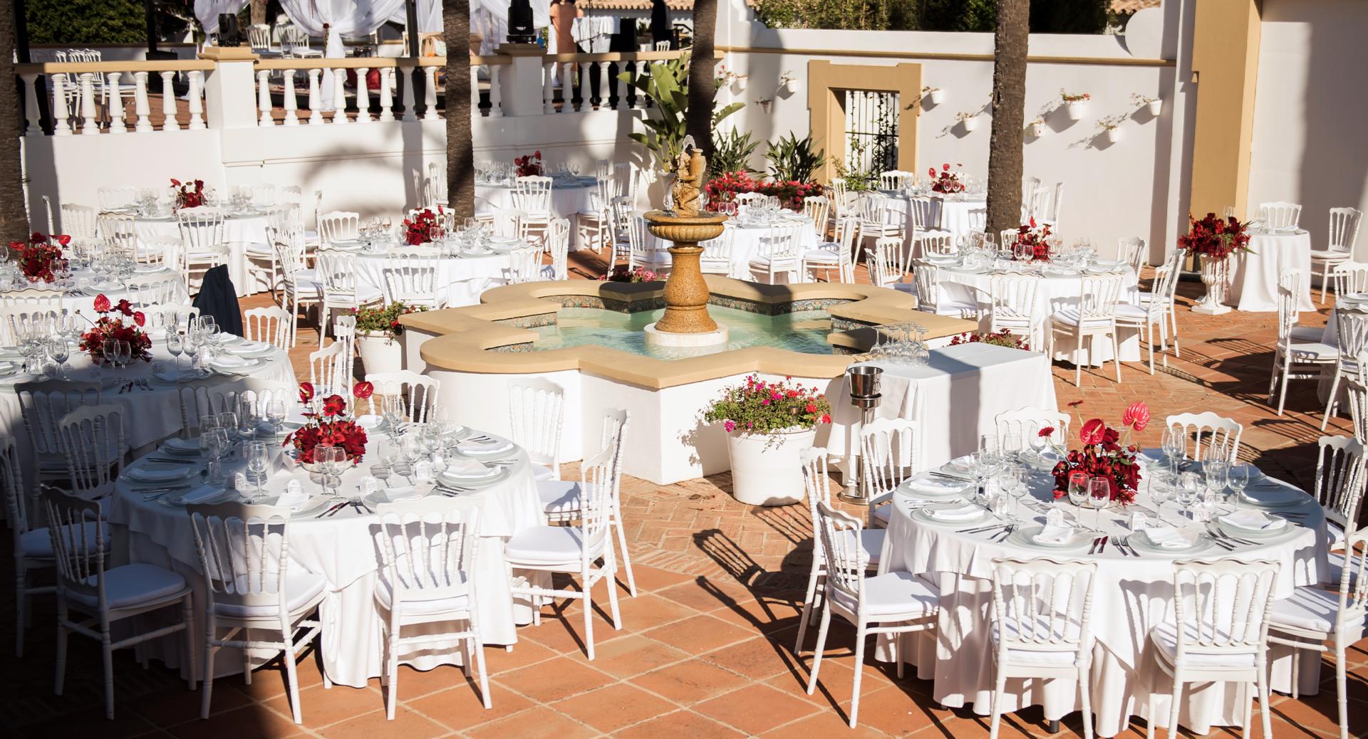 Outdoor courtyard set up for an event with round tables, white chairs and linens, red floral centerpieces, and a central decorative fountain.