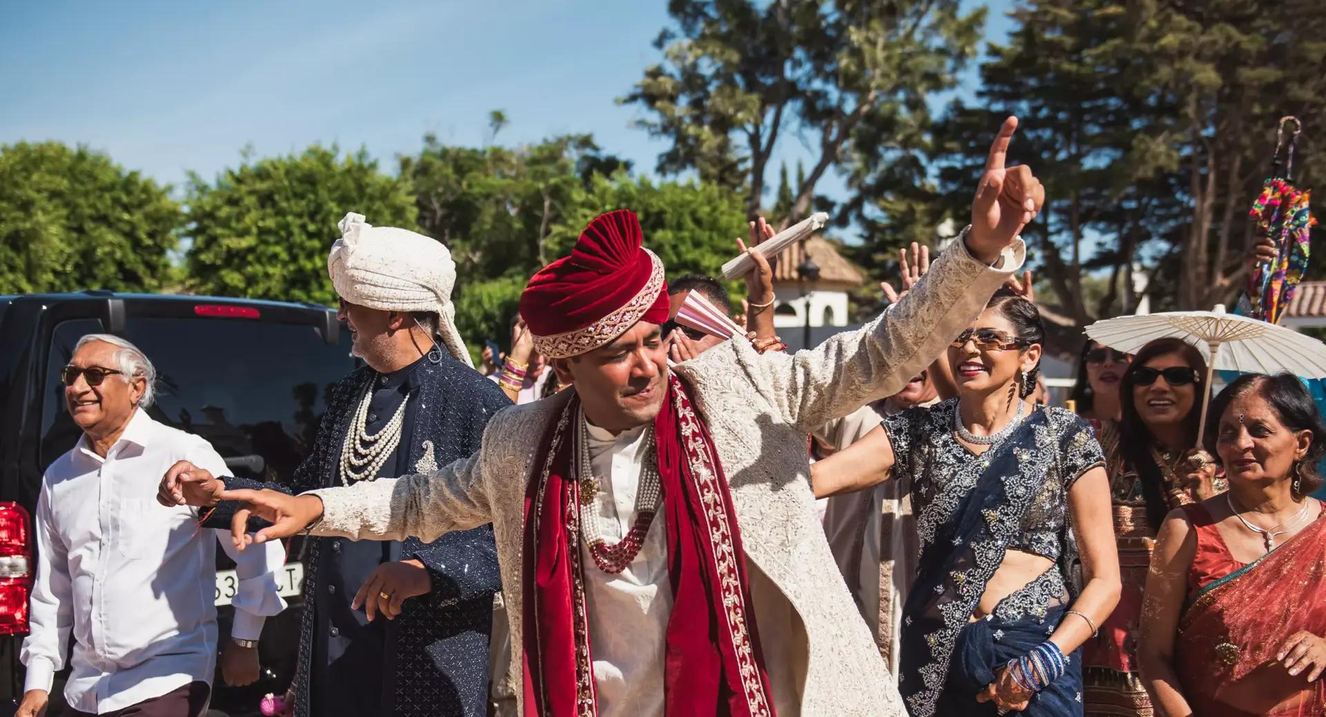 A group of people dressed in traditional Indian attire celebrate outdoors; a man in front wearing a sherwani and turban gestures energetically.