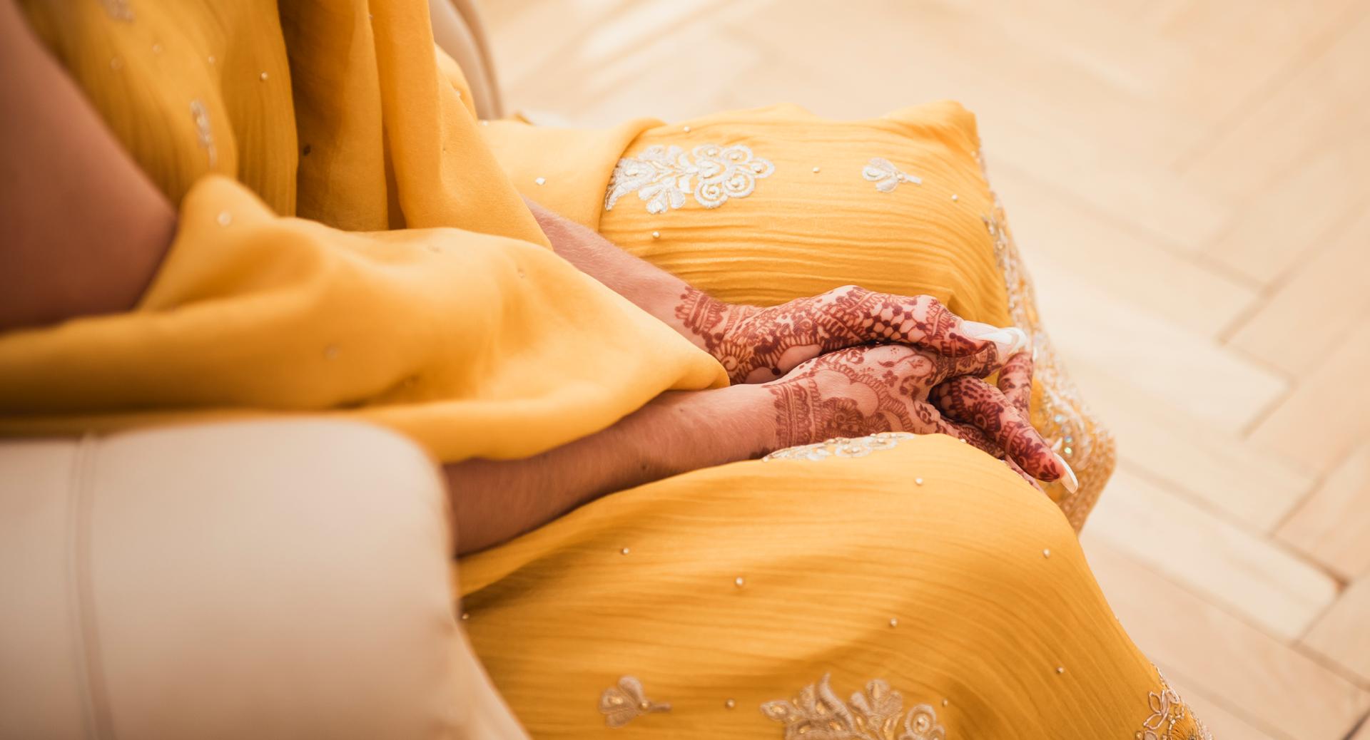 Person sitting with hands resting in lap, wearing a yellow embroidered garment and intricate henna designs on their hands.