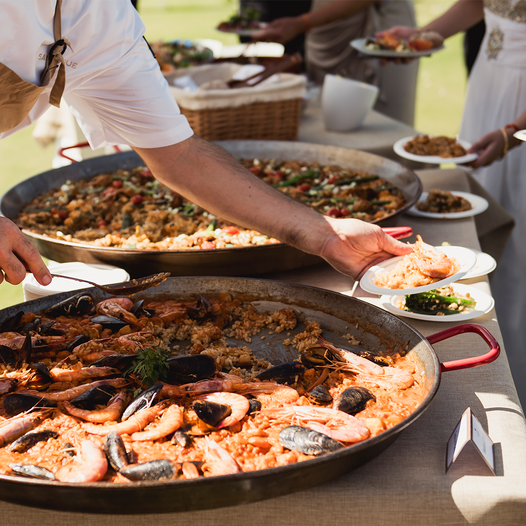 A person serves seafood paella from a large pan onto a plate at an outdoor buffet table with other dishes in the background.