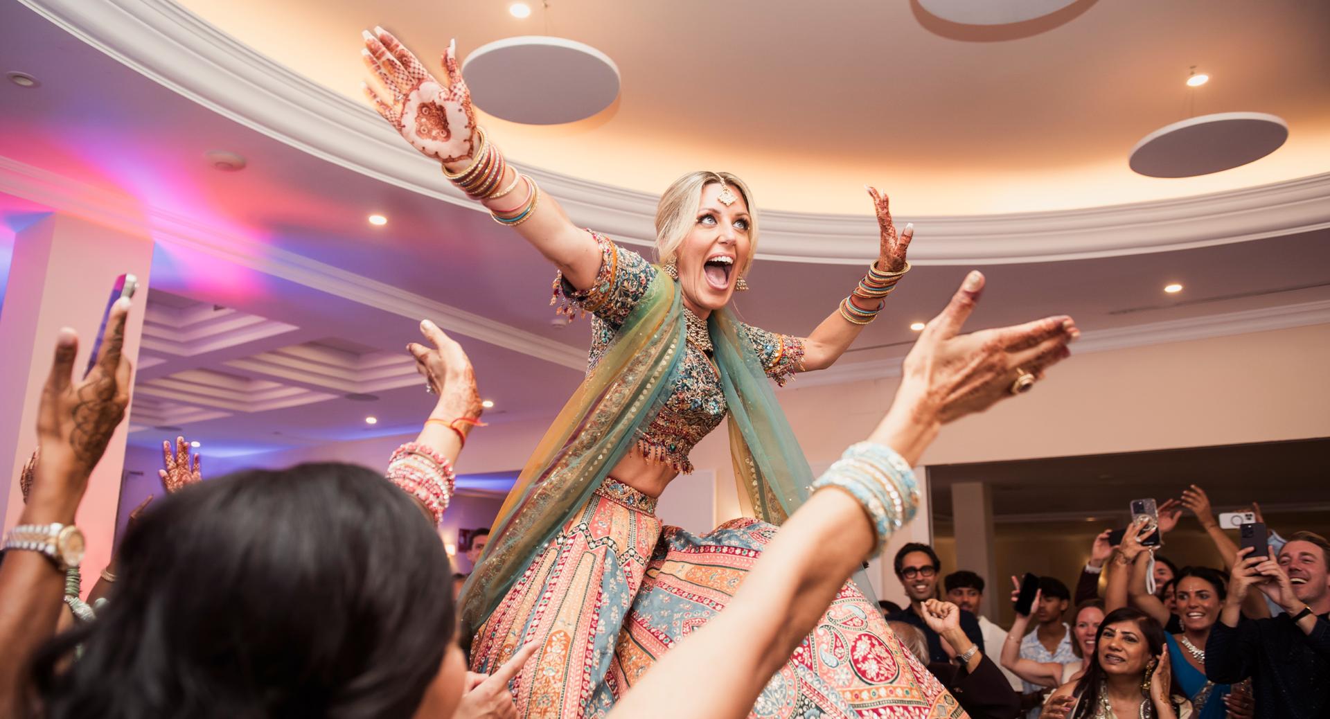 A woman in traditional Indian attire is lifted and celebrated by a crowd at an indoor event, with her arms raised and people cheering around her.