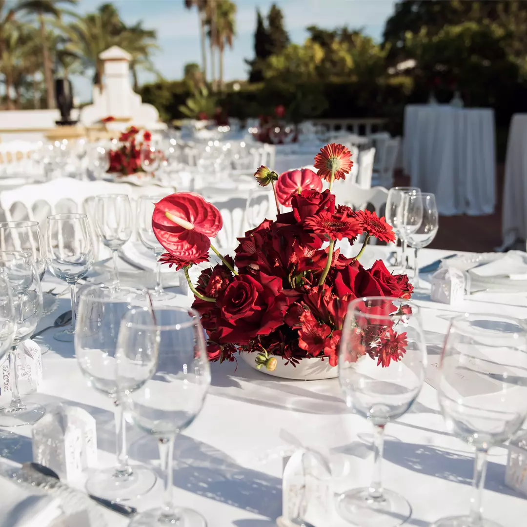 A white outdoor table is set with empty wine glasses and a centerpiece of red flowers, with more tables and greenery visible in the background.