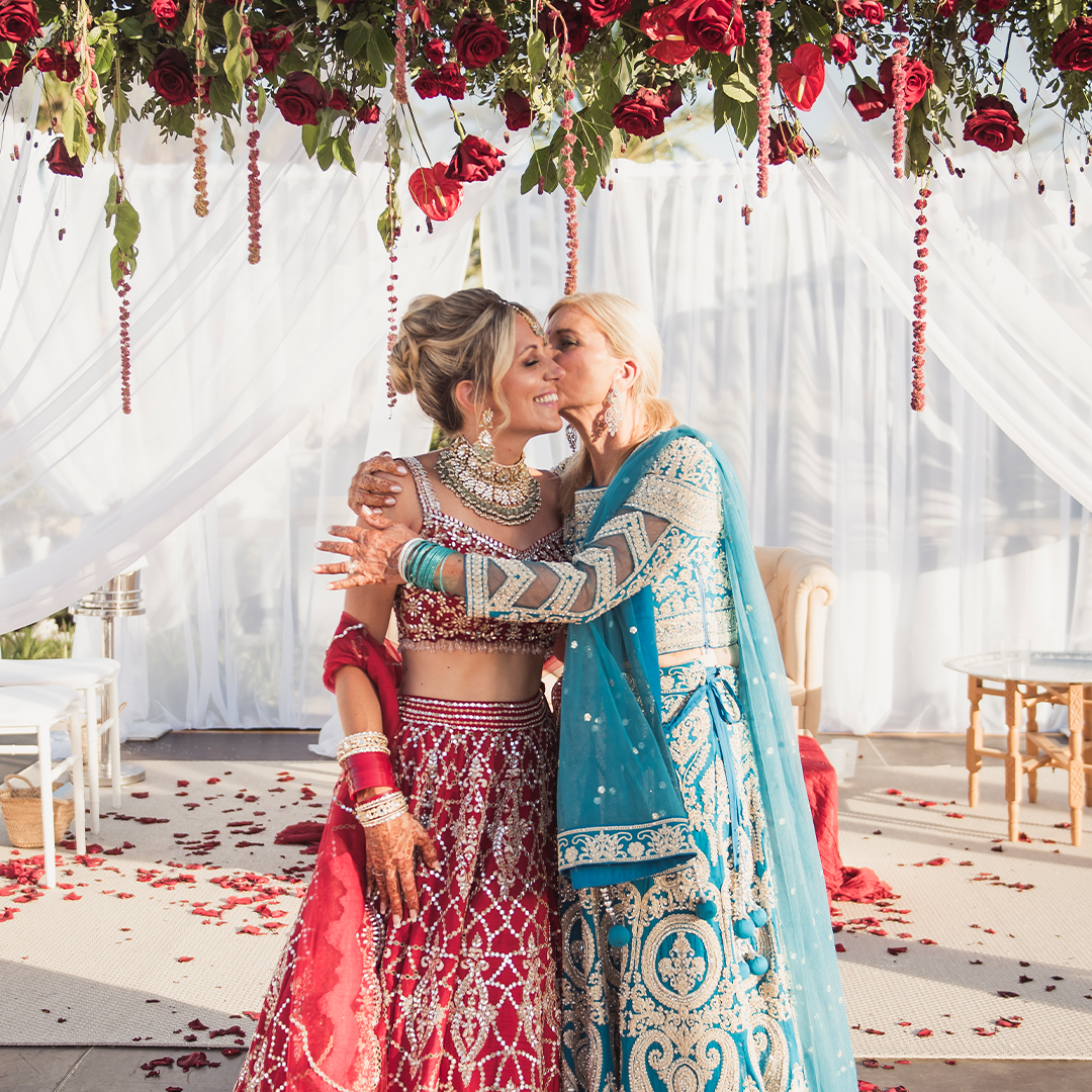Two women in ornate traditional South Asian attire embrace and exchange a kiss on the cheek under a hanging floral arrangement with red roses and white draped fabric in the background.