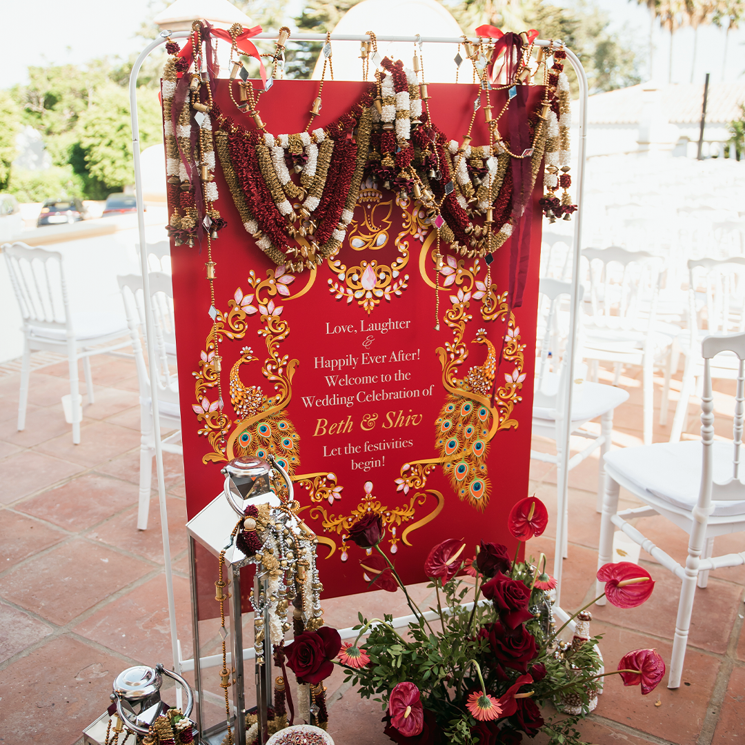 A red wedding welcome sign with floral garlands, gold designs, and decorated elephants stands among white chairs and red flowers in an outdoor setting.