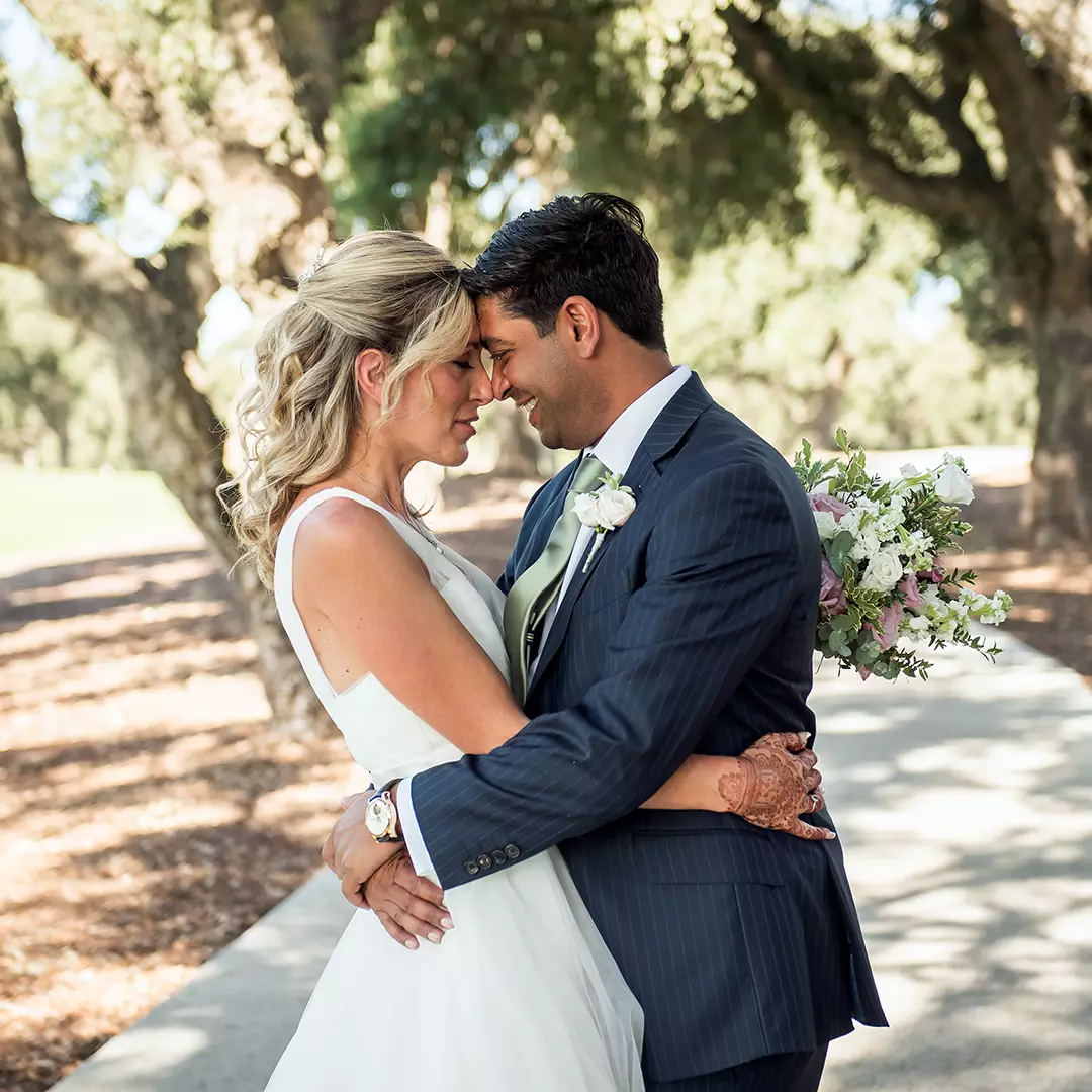 A bride and groom stand close together outdoors, embracing and touching foreheads, with trees and sunlight in the background.