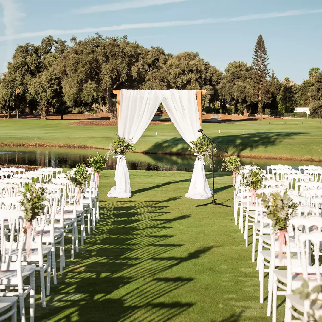 Rows of white chairs face a wedding arch with white drapes and floral arrangements, set outdoors on a grassy area near trees and a pond.