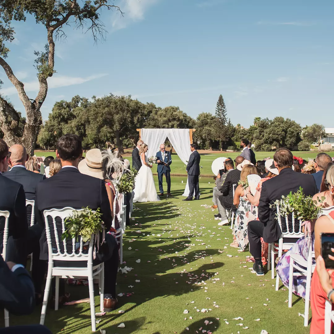 Outdoor wedding ceremony with guests seated on either side of an aisle, bride and groom standing under a white arch, trees and blue sky in the background.