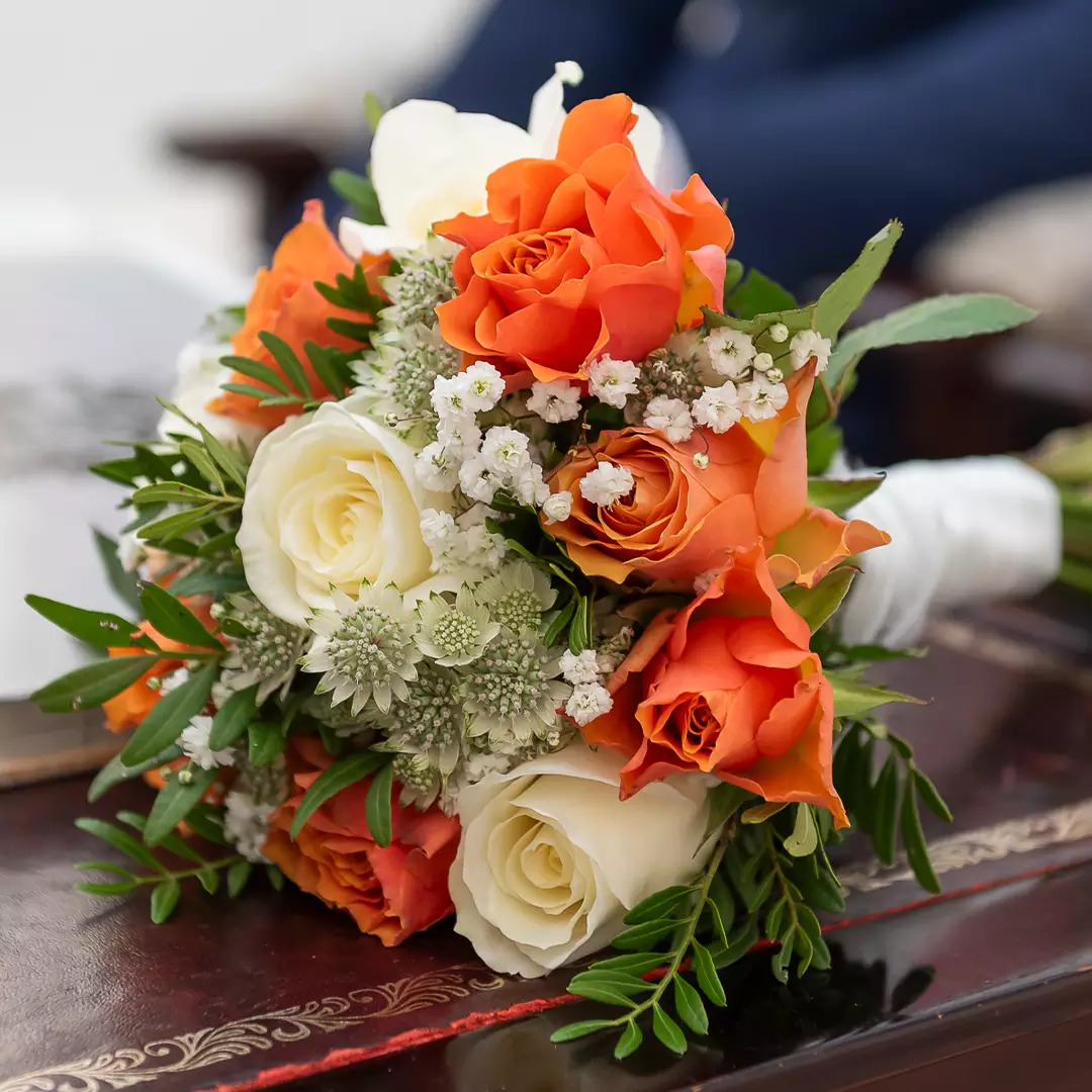 A bouquet of orange and white roses, accented with green leaves and small white filler flowers, rests on a dark wooden surface.