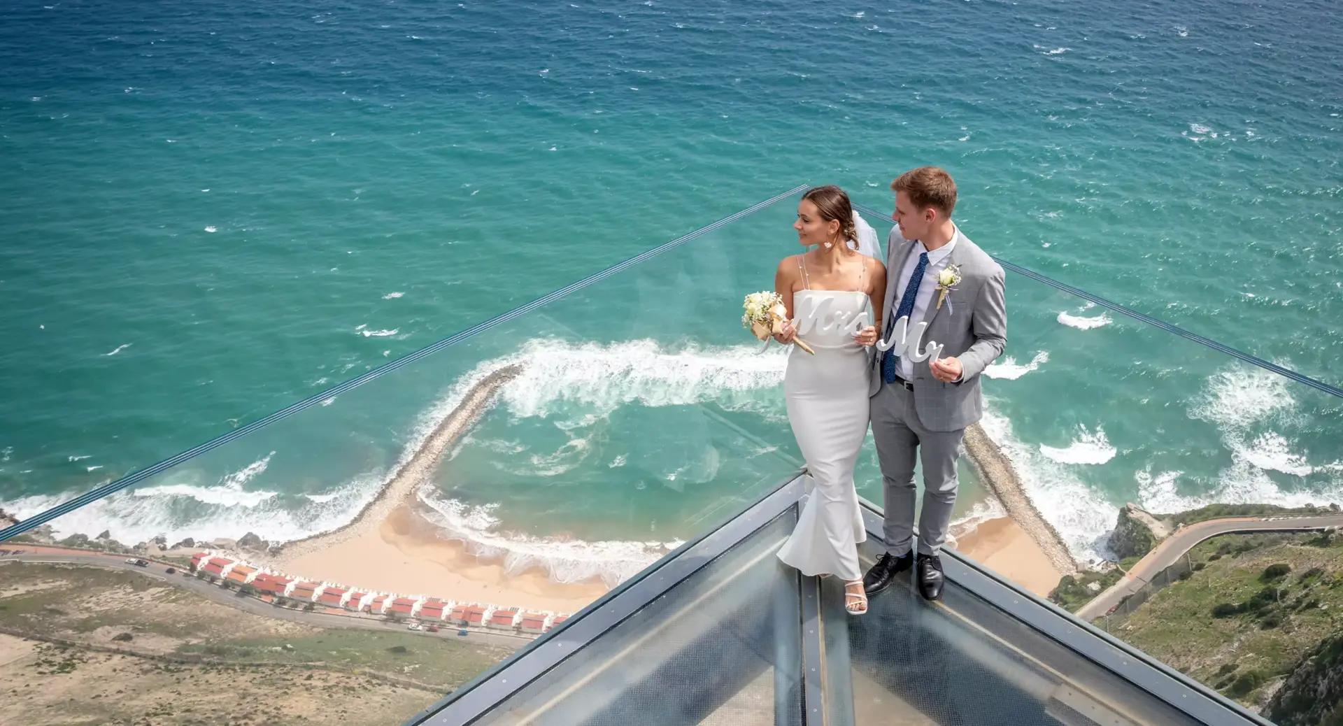 A bride and groom stand on a glass platform high above a coastal landscape, with waves and a beach visible far below.