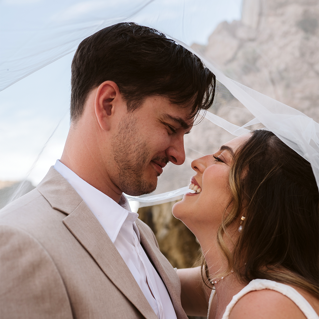 A couple dressed in wedding attire stands close together outdoors, smiling beneath a sheer veil.