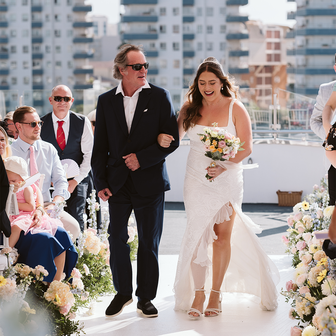 A bride in a white dress walks down an outdoor aisle with an older man, surrounded by seated guests and floral arrangements, with city buildings in the background.