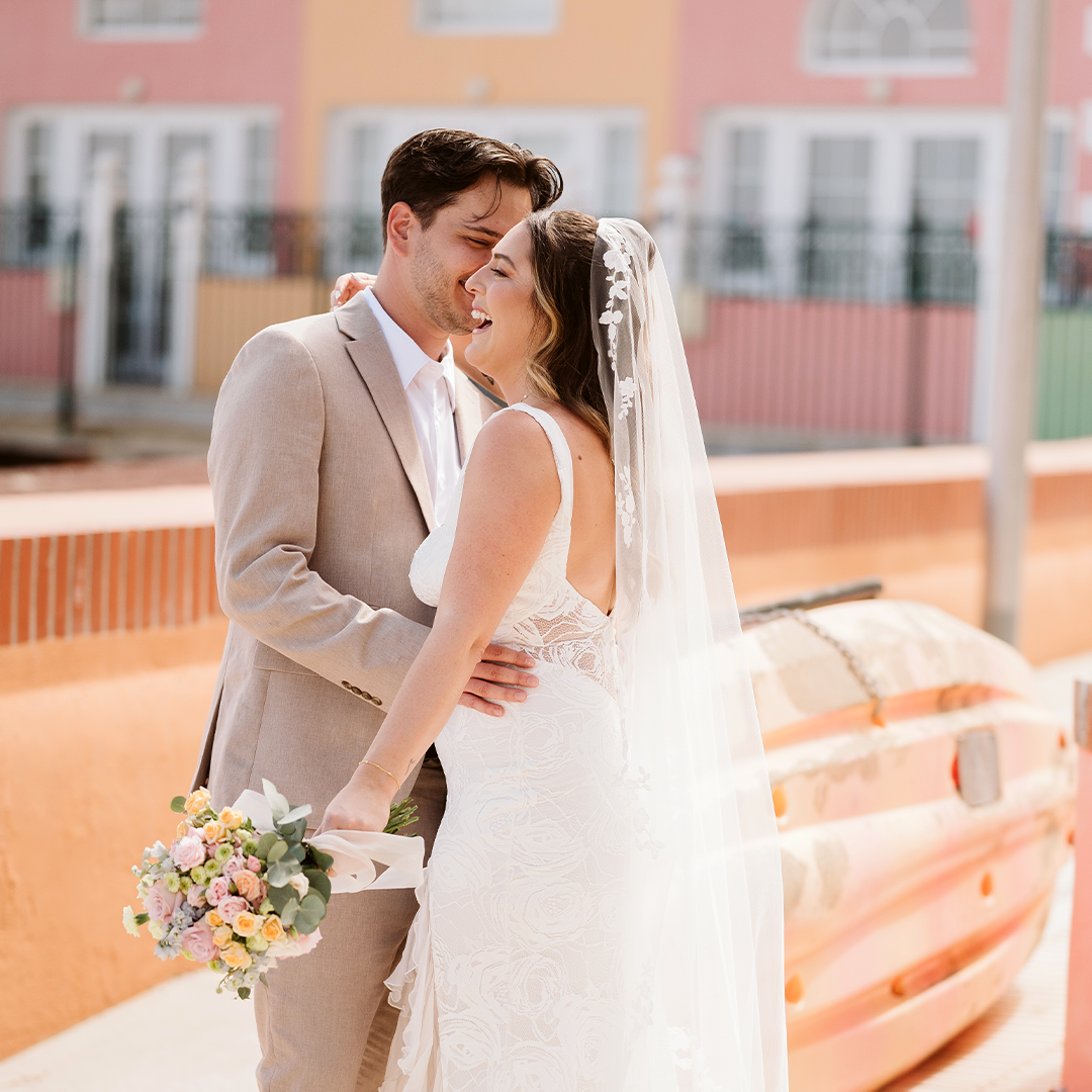 A bride and groom stand close together outdoors, smiling. The bride holds a bouquet and wears a lace dress and veil; colorful buildings and a boat are in the background.