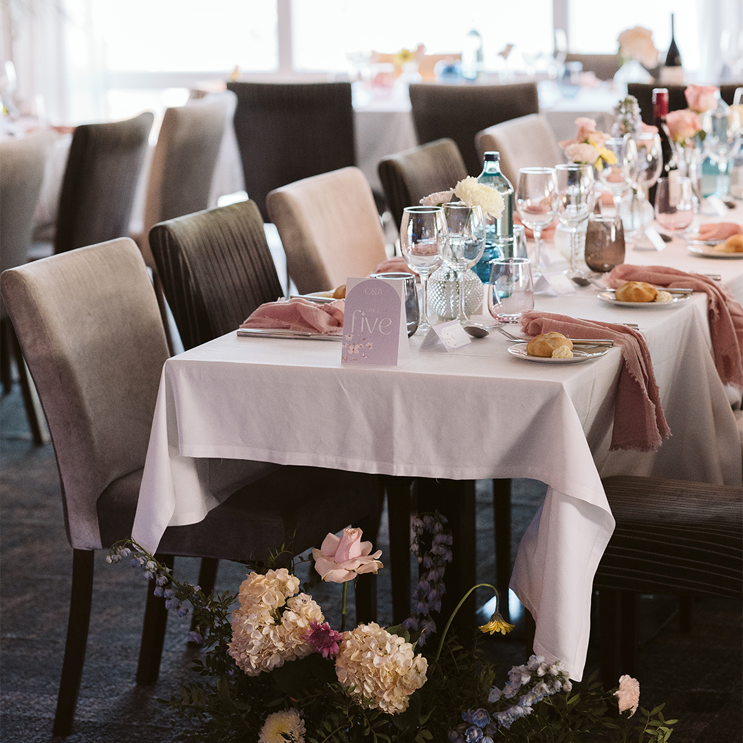 Elegant dining table set for an event with glassware, plates, bread rolls, pink napkins, a table number card, and flower arrangements.