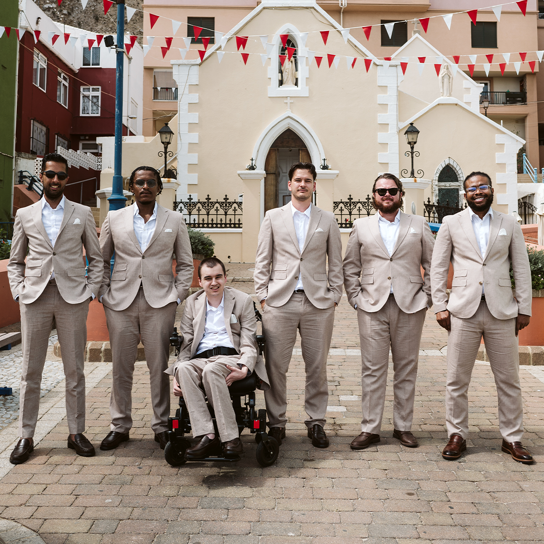 Six men in beige suits, one seated in a wheelchair, pose together outside in front of a cream-colored building decorated with red and white bunting.