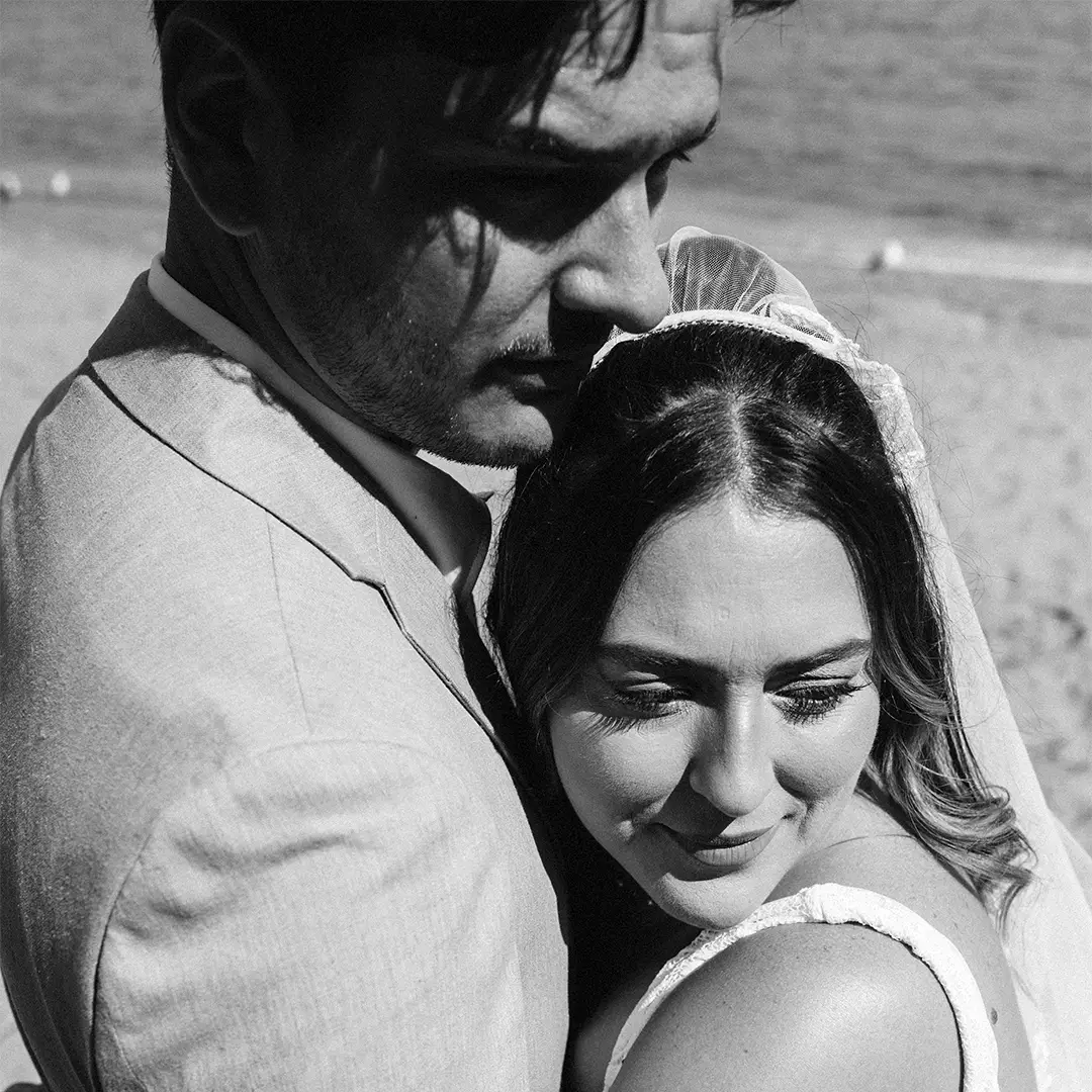 A bride and groom embrace on a beach, with the groom looking away and the bride smiling softly, captured in black and white.