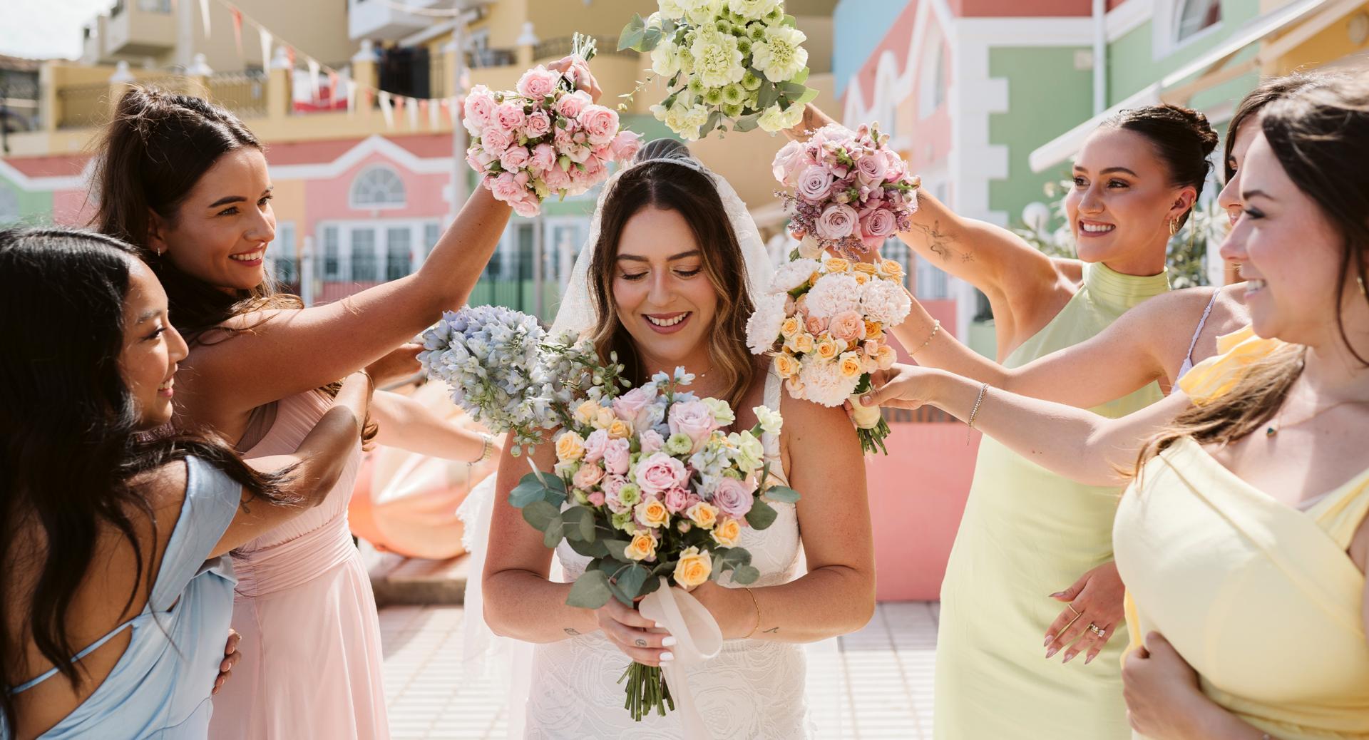 A bride in a white dress holds a bouquet while bridesmaids in pastel dresses surround her with flowers in an outdoor setting.