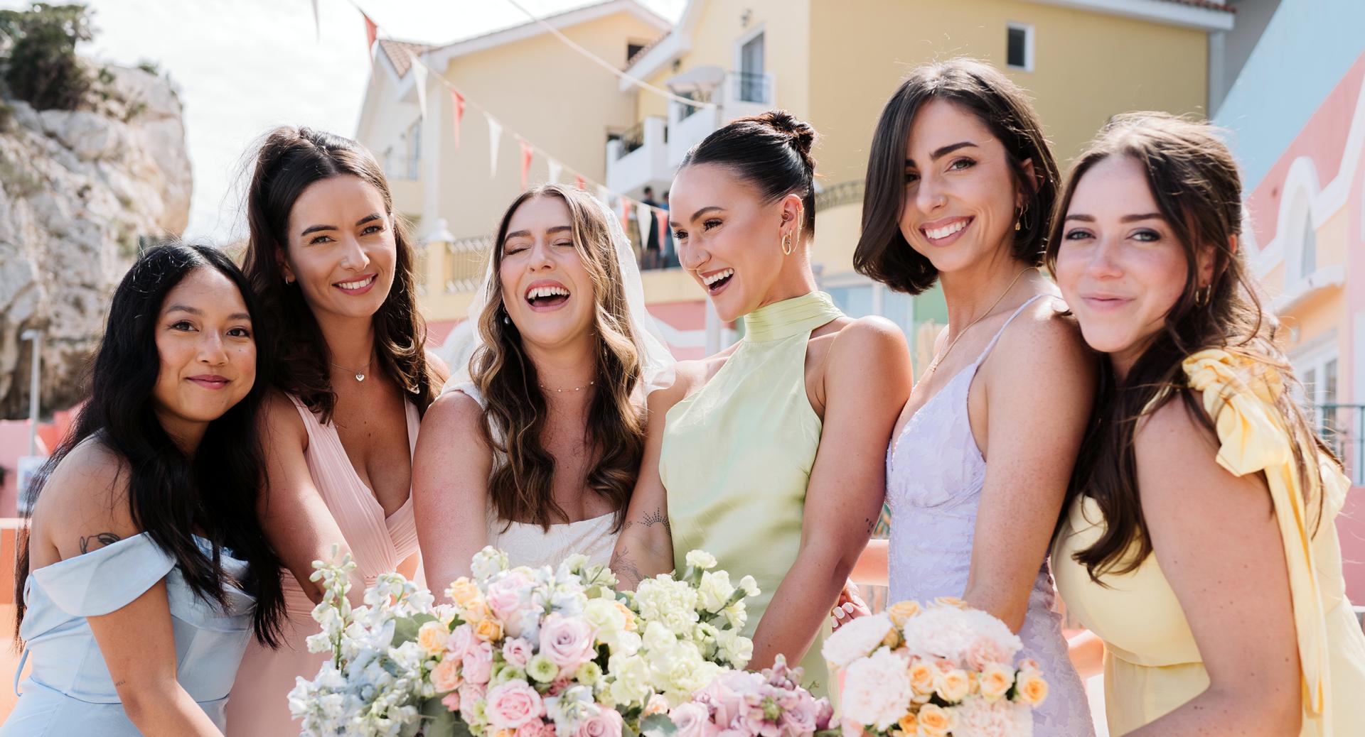Six women in colorful dresses stand close together outdoors, smiling and holding floral bouquets. Bright buildings and decorations are visible in the background.