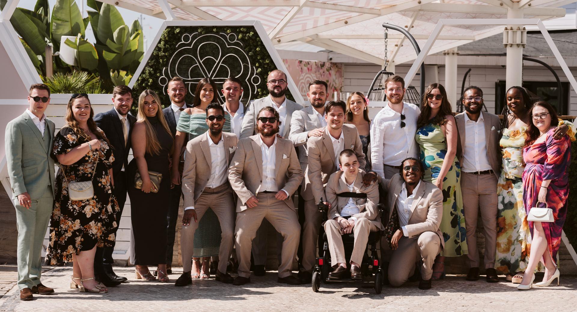 A group of people dressed in formal attire pose together outdoors in front of a floral and geometric backdrop on a sunny day.