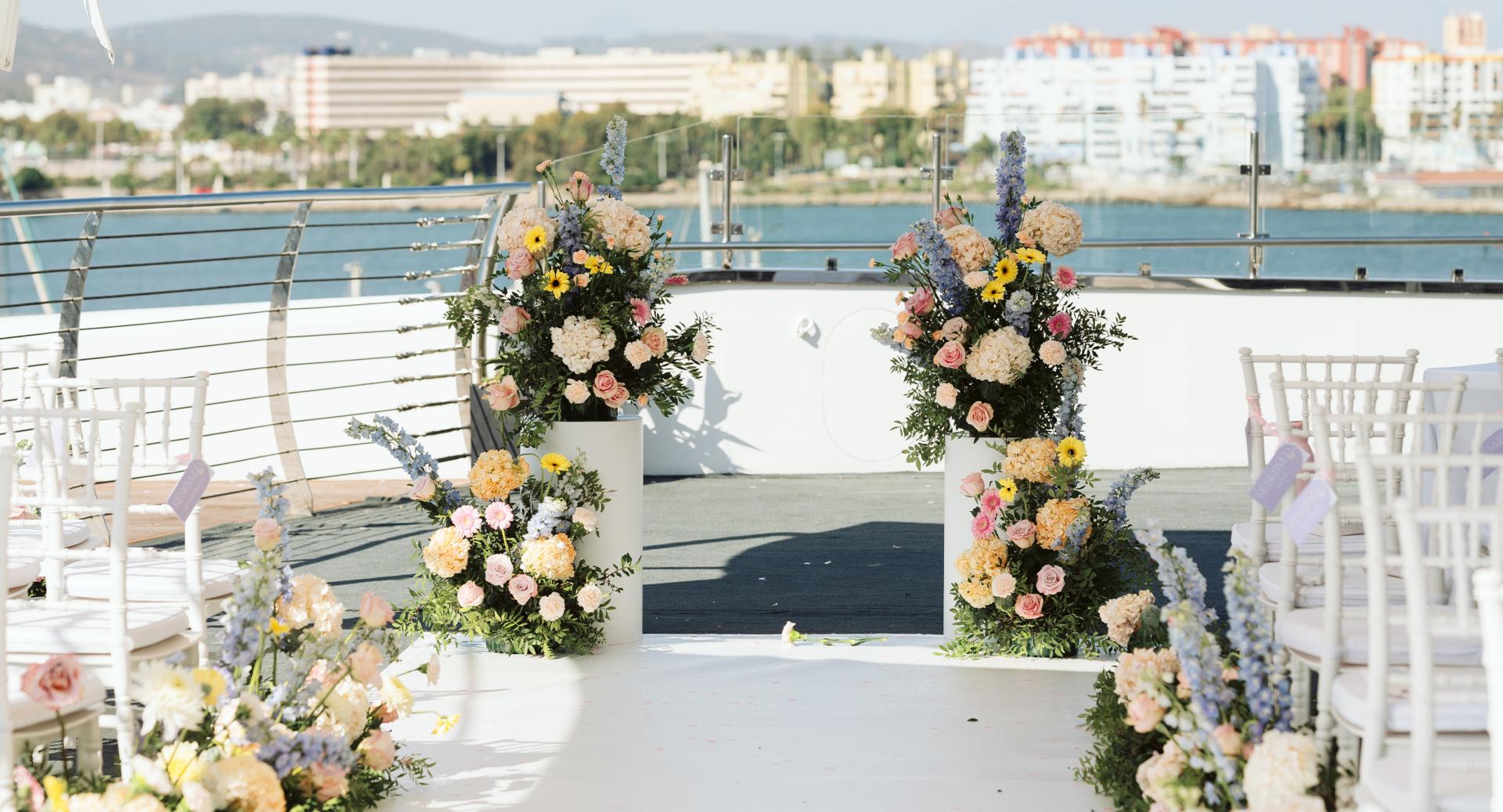 An outdoor wedding ceremony setup with white chairs and large floral arrangements on a deck overlooking water and city buildings in the background.