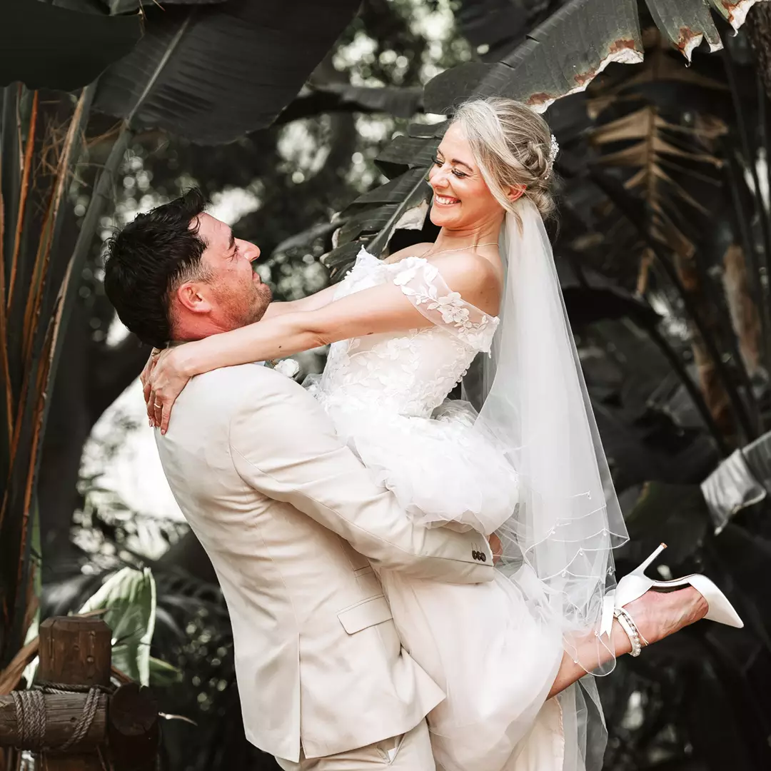 A groom in a light suit lifts a smiling bride in a white wedding dress and veil outdoors, surrounded by lush green foliage.