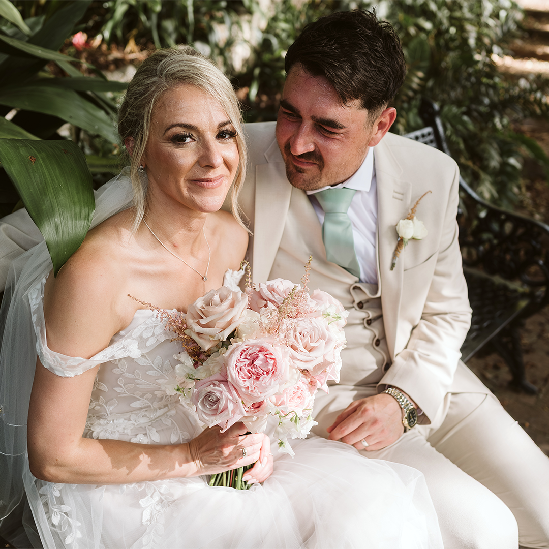 A bride in a white dress holds a bouquet of pink and white flowers while sitting next to a groom in a beige suit on an outdoor bench.