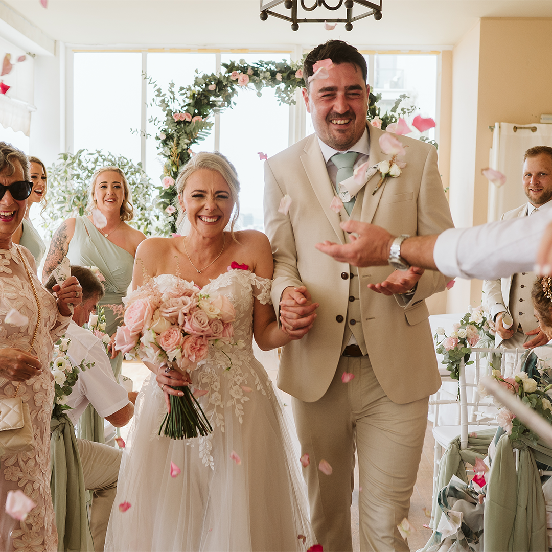 A bride and groom walk down the aisle smiling, surrounded by guests throwing rose petals in a brightly lit room decorated with flowers.