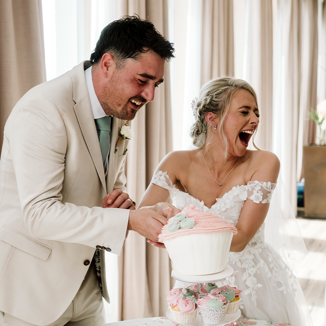 A bride and groom in formal attire laugh while cutting a large cupcake-shaped wedding cake together indoors.