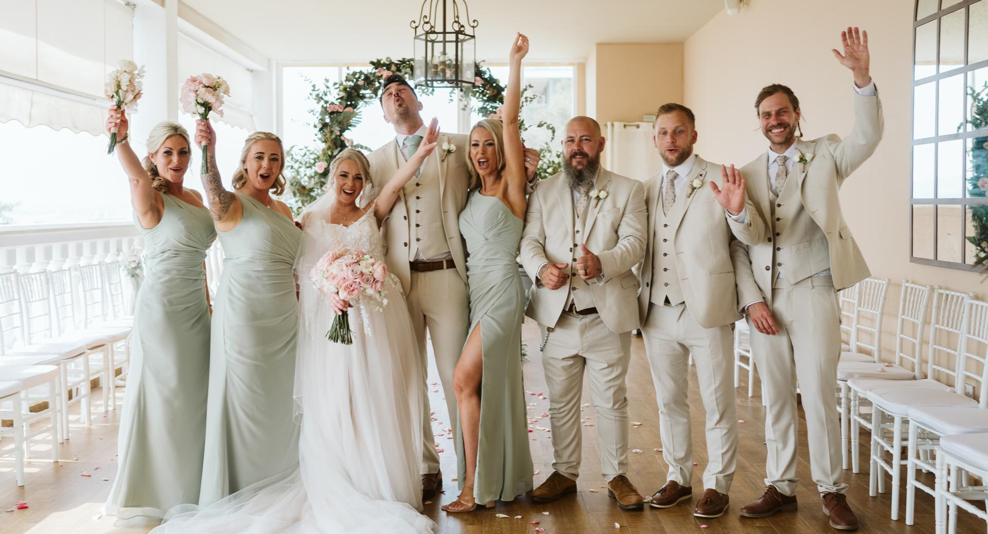 A bride and groom stand with bridesmaids and groomsmen in light green and beige outfits, smiling and raising their arms in celebration in a bright wedding venue.