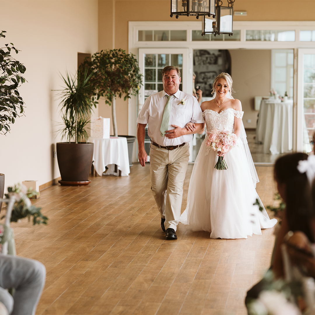 A bride in a white gown holding a bouquet walks down the aisle with a man in a white shirt and tie in a bright indoor venue.