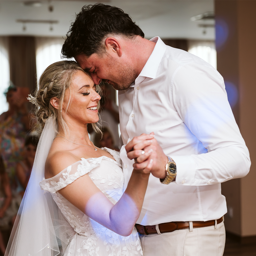 A bride and groom in white attire share a close moment while dancing together indoors, smiling and holding hands.