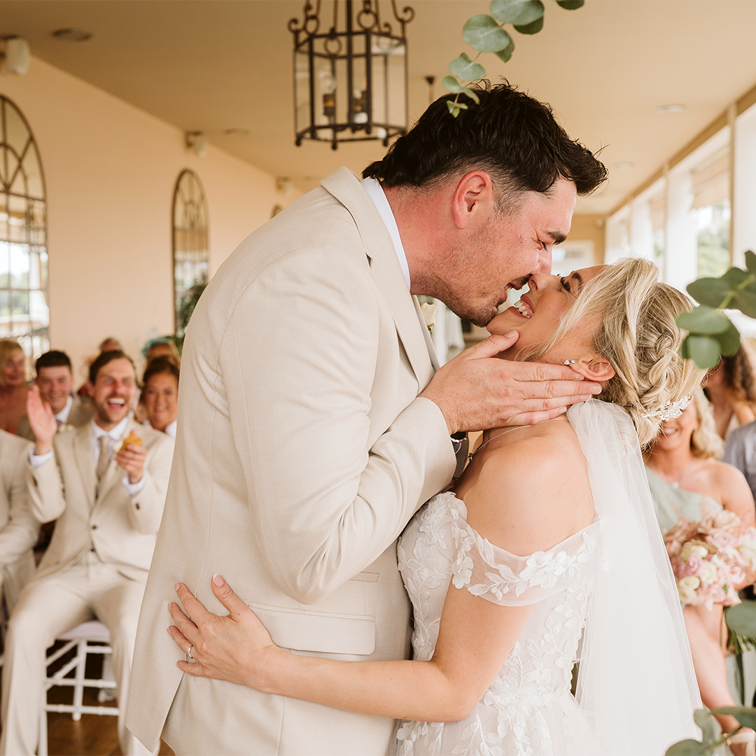 A bride and groom share a kiss at their wedding ceremony, surrounded by seated guests who are smiling and clapping.