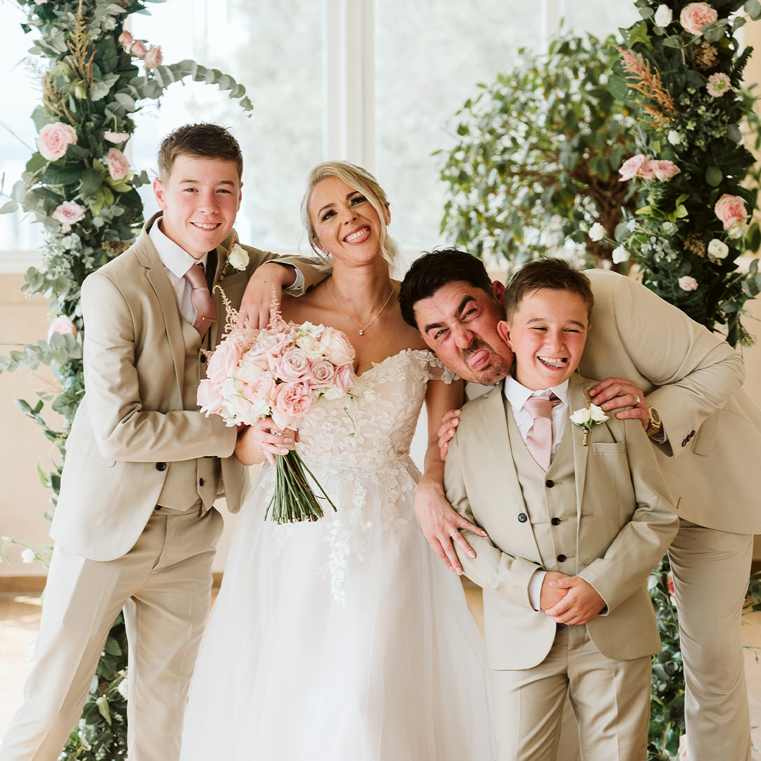 A bride in a white dress holds a bouquet and poses playfully with three men in beige suits under a floral arch.