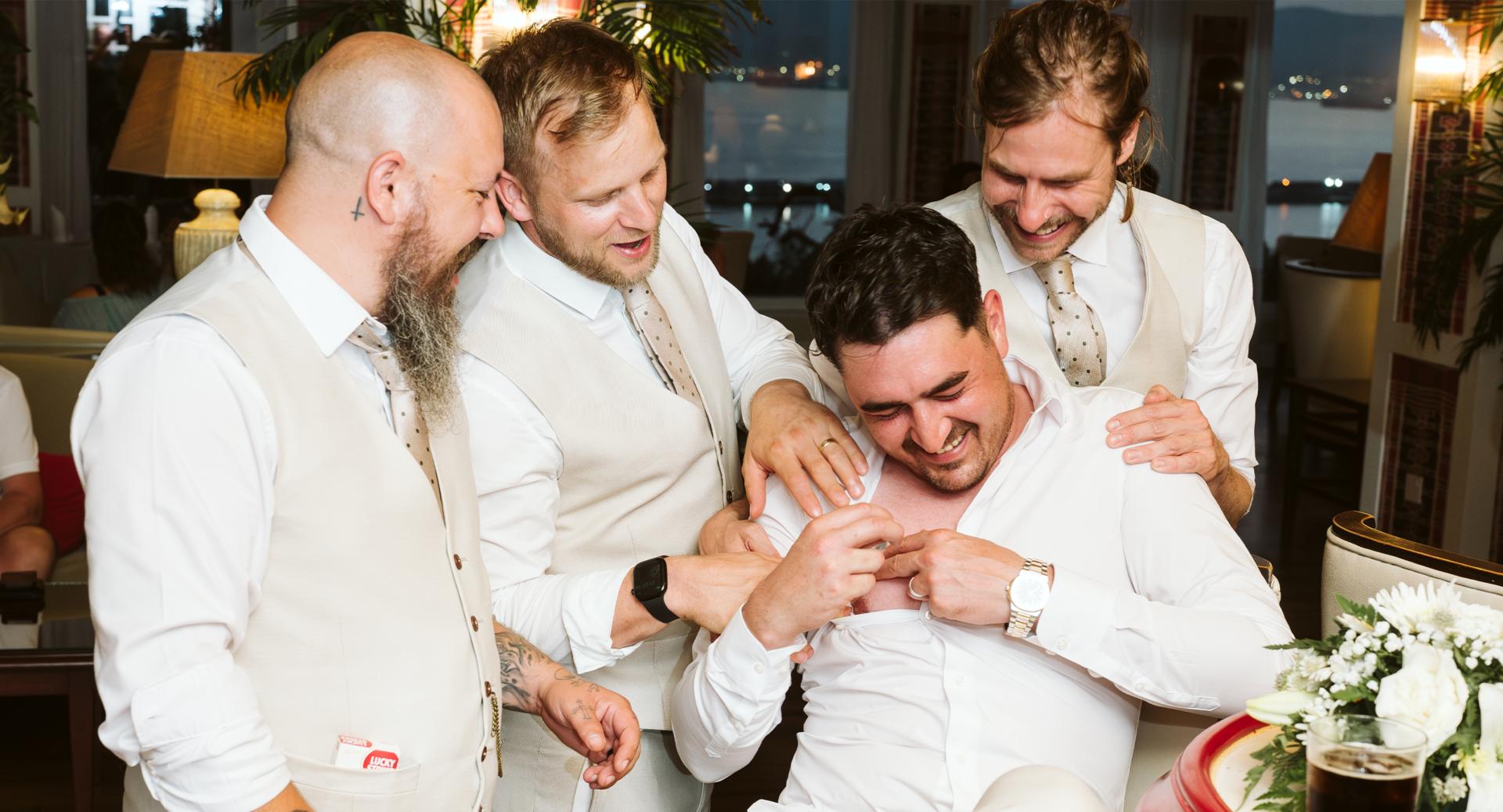 Four men dressed in light-colored suits laugh and playfully interact indoors, with one man seated as the others gather around him. A floral arrangement is visible in the foreground.