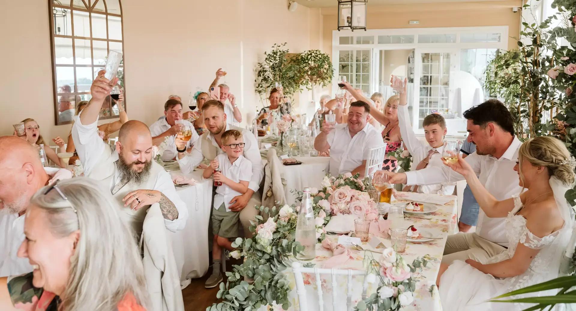 A group of people sit around decorated tables at a wedding reception, raising their glasses in a toast.